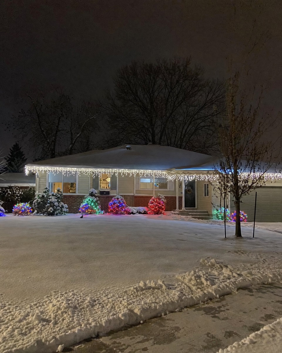 Snowy ranch home with white icicle lights along the whole roofline and multicolor shrubs