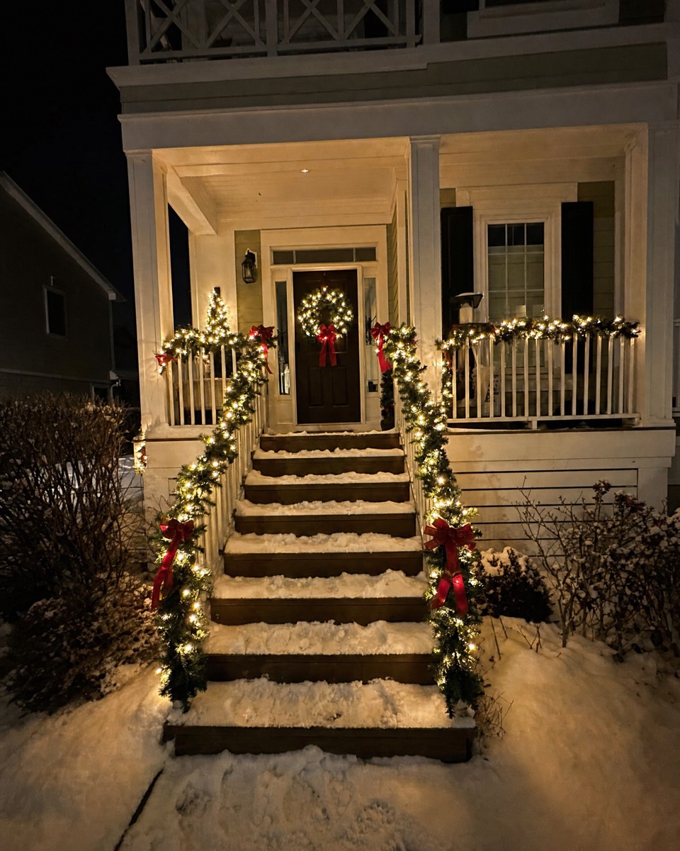Snowy front steps with railings wrapped in lit garland and red bows